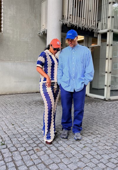 Dos personas están juntas: una lleva un conjunto de punto de un patrón azul y crema, mientras que la otra lleva una camisa a rayas azul y pantalones azul real.