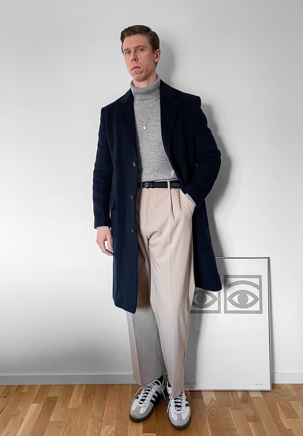 Giovane uomo con cappotto blu navy, dolcevita grigio, pantaloni beige e sneakers in piedi contro un muro bianco con un'opera d'arte raffigurante un occhio incorniciata sul pavimento.