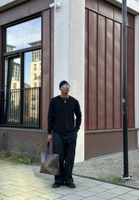 Homem em traje preto e chapéu está na calçada da cidade segurando uma grande bolsa tote de textura marrom perto da esquina de um edifício.
