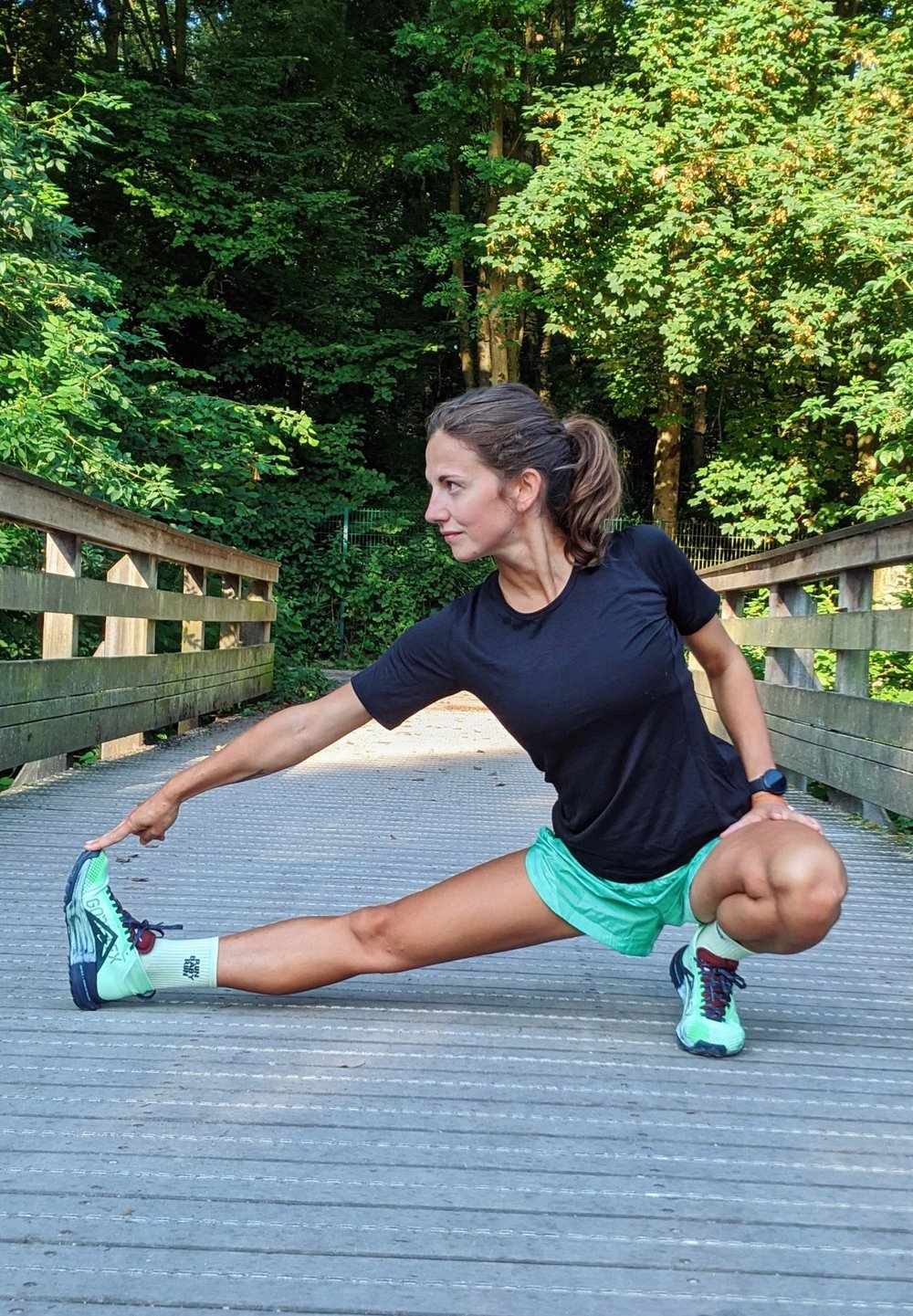 Mulher atlética a alongar-se numa ponte de madeira, vestindo uma t-shirt preta, calções verdes e ténis de corrida multicoloridos com detalhes brancos.