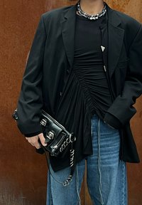 Blazer oversized preto sobre um top preto franzido, combinado com jeans azul claro. Uma bolsa preta com hardware prateado e detalhe em corrente é segurada.