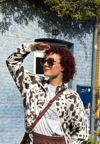 Vit jacka med svart ko-mönster, vit t-shirt, bruna byxor, överdimensionerade solglasögon, står mot en blå vägg med grönska ovanför.