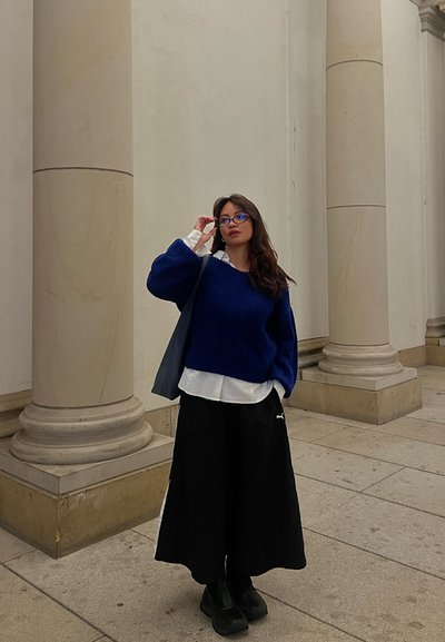 Mujer con suéter azul, camisa blanca, pantalones anchos negros y gafas, de pie entre columnas de piedra en un pasillo de un edificio.
