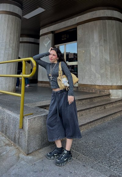 Top gris de manga larga, pantalones cortos amplios negros y zapatillas negras. Bolsa amarilla con un accesorio de peluche. Fondo de escalones de hormigón y barandilla amarilla.