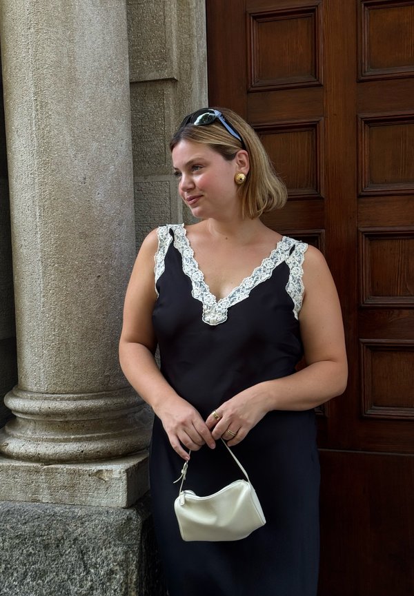 Donna con abito nero con rifiniture in pizzo bianco che tiene una piccola borsa bianca, in piedi accanto a una colonna di pietra e a una porta in legno con pannelli.