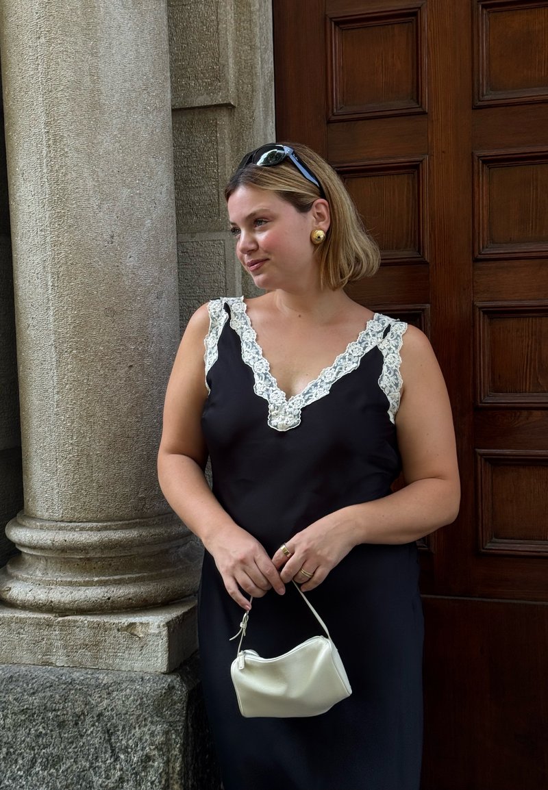 Femme en robe noire ornée de dentelle blanche tenant un petit sac blanc, debout près d'une colonne en pierre et d'une porte en bois à panneaux.