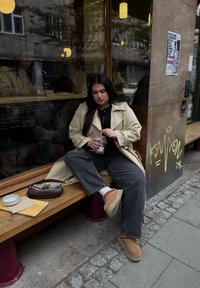 Bež trench coat čez črn top, temne ohlapne traperke, svetlo rjave natikače, z rjavim ročajem. Pijača je v desni roki.