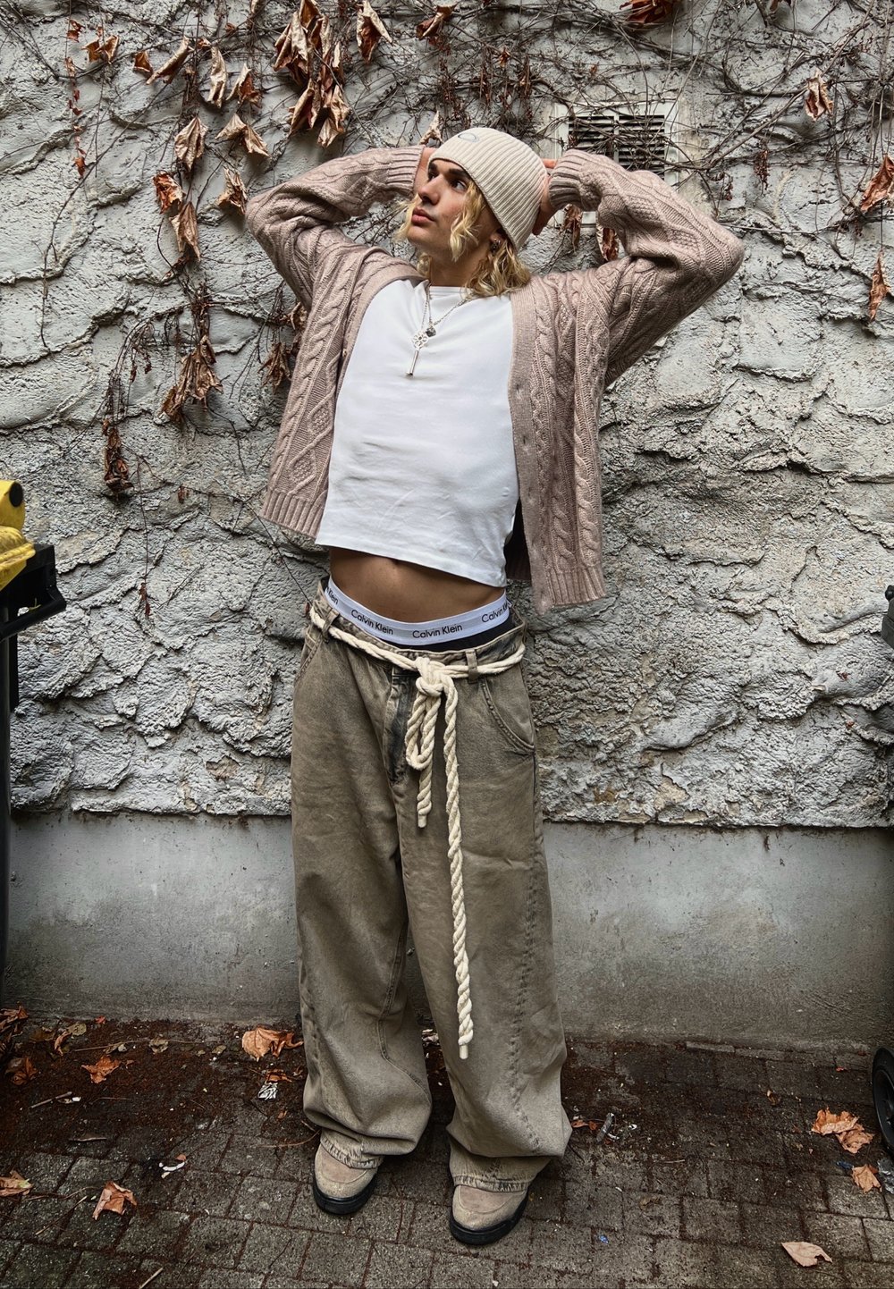 Cardigan beige lavorato a maglia, camicia bianca corta, pantaloni beige larghi con cintura a corda e scarpe beige. Sullo sfondo si osserva un muro grigio testurizzato e foglie secche.
