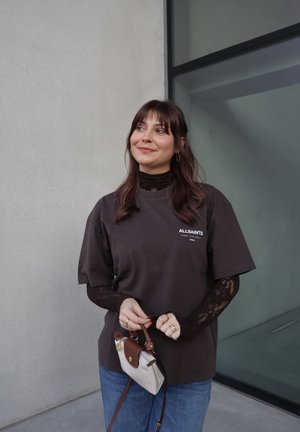 Mujer joven de cabello oscuro con una camiseta negra de AllSaints sobre una blusa de encaje negra, jeans azules, sosteniendo un bolso pequeño beige y marrón.