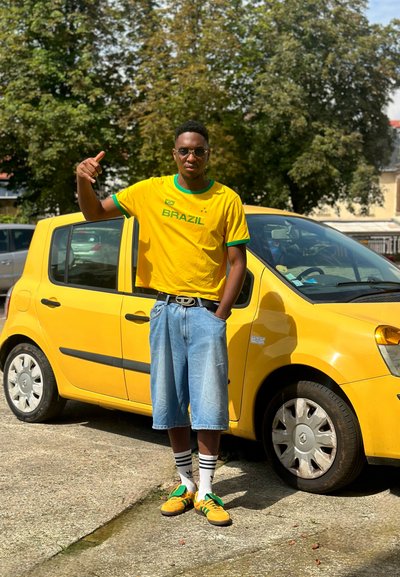 Camiseta amarilla de Brasil con detalles verdes, pantalones cortos de mezclilla holgados, zapatillas amarillas y verdes, calcetines blancos con rayas negras, junto a un coche amarillo.