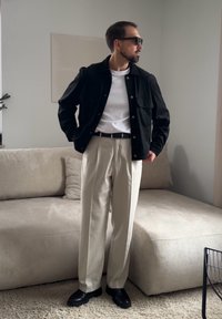 Άνδρας που φοράει μαύρο μπουφάν, λευκό πουκάμισο, μπεζ πιετέ παντελόνι, μαύρη ζώνη και μαύρα παπούτσια, στέκεται σε ένα σαλόνι με ουδέτερους τόνους.