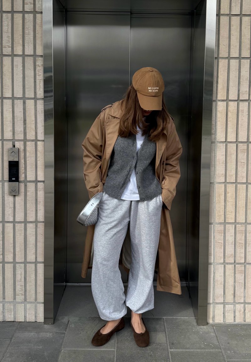 Persona che indossa un cappellino marrone con testo, trench beige, gilet grigio, camicia bianca, pantaloni tuta grigi e ballerine marroni, in piedi in un ascensore chiuso.