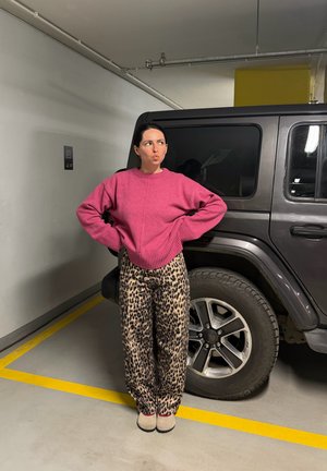 Mujer con suéter rosa y pantalones de estampado de leopardo, de pie con las manos en las caderas junto a un SUV negro en un estacionamiento subterráneo.