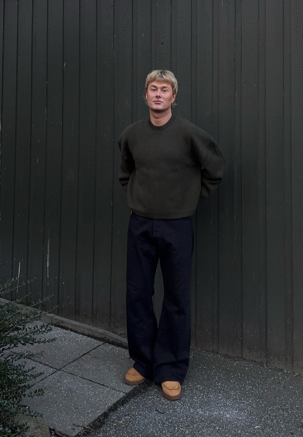 Pull vert foncé avec un col côtelé, associé à un pantalon noir ample et des chaussures à enfiler couleur fauve, sur fond d'un mur en bois foncé.