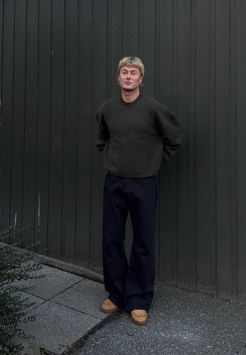 Pull vert foncé avec un col côtelé, associé à un pantalon noir ample et des chaussures à enfiler couleur tan, sur fond d'un mur en bois sombre.