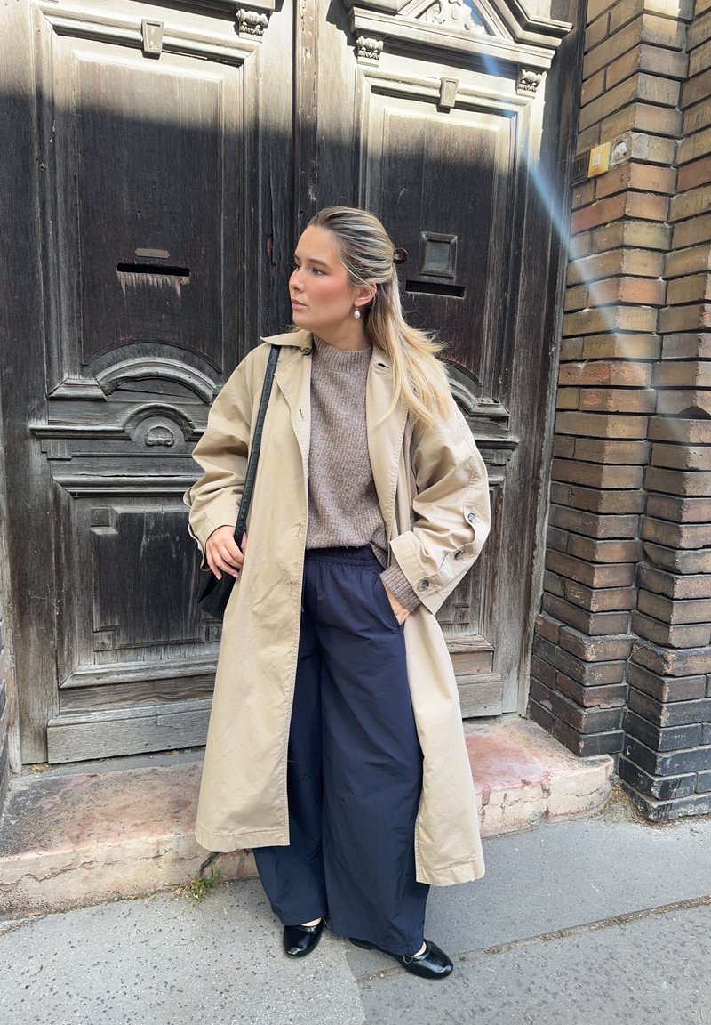 Casaco trench bege sobre um suéter castanho-claro, combinado com calças largas azul-marinho e sapatos pretos. Design simples, textura suave e ajuste casual.