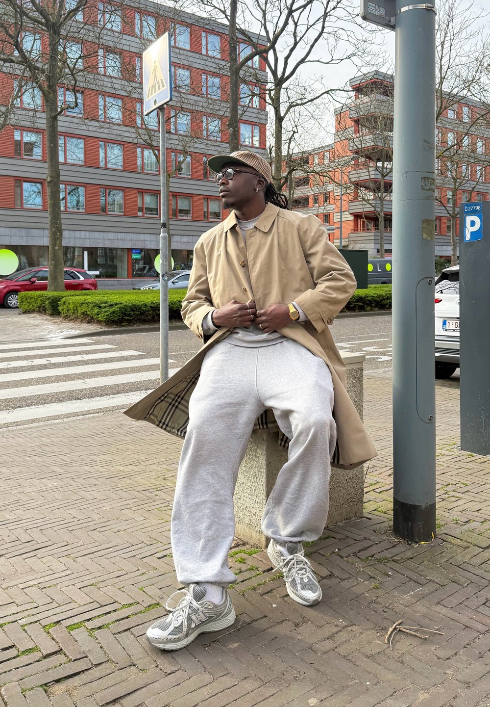 Beige trenchcoat över en grå sweatshirt, grå sweatpants och grå sneakers med detaljer i nät. Urban miljö med träd och byggnader.