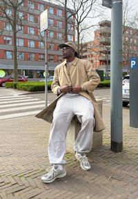 Beige trenchcoat över en grå sweatshirt, grå sweatpants och grå sneakers med detaljer i nät. Urban miljö med träd och byggnader.