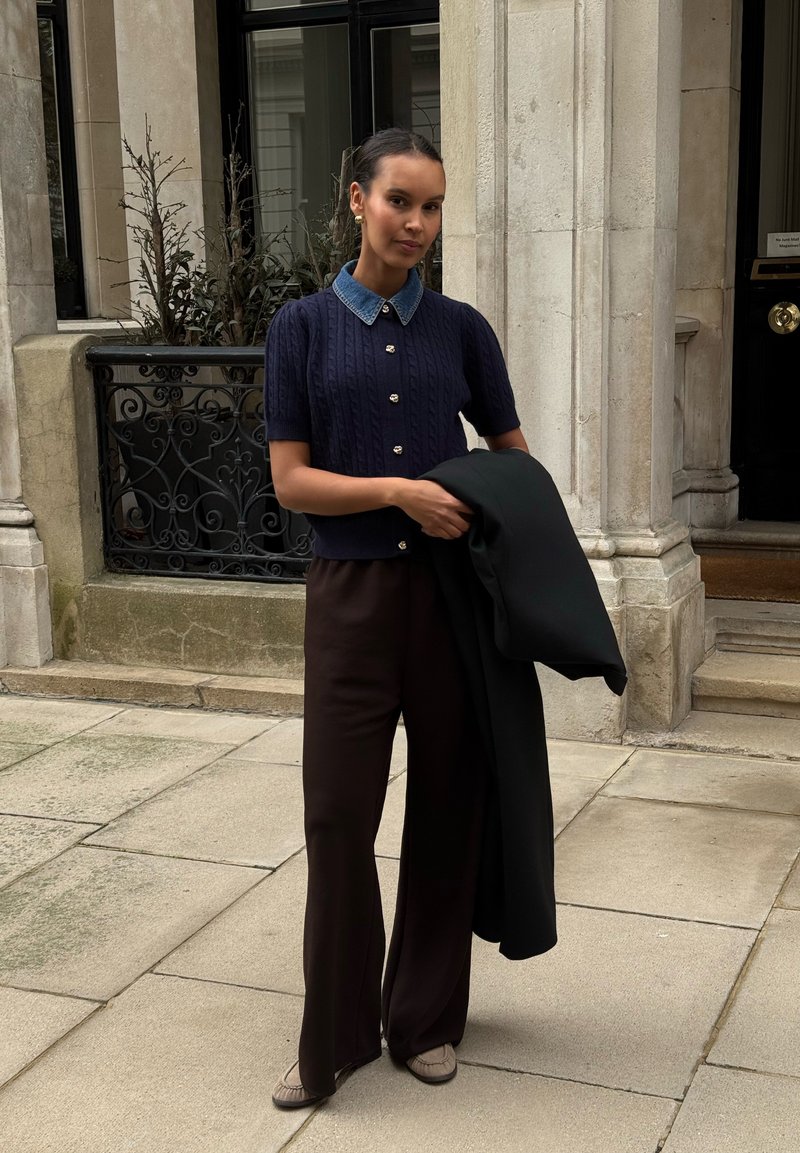 Marineblauer Pullover mit Kurzarm und Zopfmuster, einem Jeanskragen, braune Hosen mit weitem Bein, hält einen schwarzen Mantel, steht auf Steinpflaster.