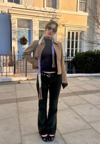 Veste courte en daim marron portée sur un col roulé noir, associée à un pantalon évasé en velours côtelé foncé et des chaussures vernies noires avec des nœuds. Longue écharpe multicolore.