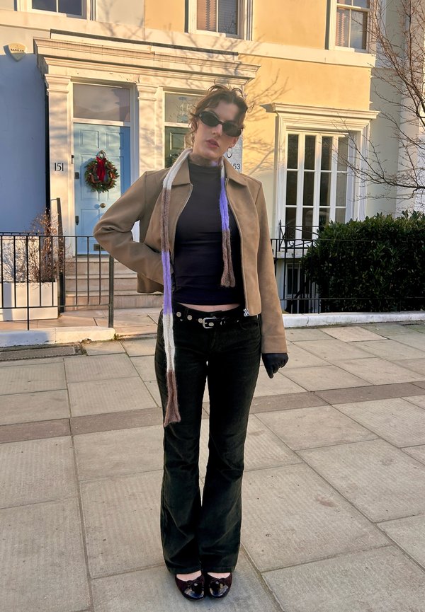 Veste courte en daim marron portée sur un col roulé noir, associée à un pantalon évasé en velours côtelé foncé et des chaussures vernies noires avec des nœuds. Longue écharpe multicolore.