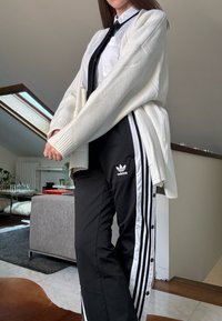 Hvit strikket cardigan lagd over en hvit skjorte med svart slips, kombinert med svarte Adidas joggebukser med hvite side-striper og knapper.
