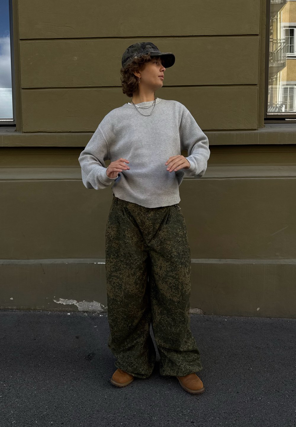 Helles graues, geripptes Sweatshirt, lockere grüne gemusterte Hosen, braune Stiefel und eine schwarze Mütze mit Ösen, vor einer gedämpften grünen Wand.