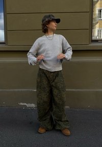 Helles graues, geripptes Sweatshirt, lockere grüne gemusterte Hosen, braune Stiefel und eine schwarze Mütze mit Ösen, vor einer gedämpften grünen Wand.