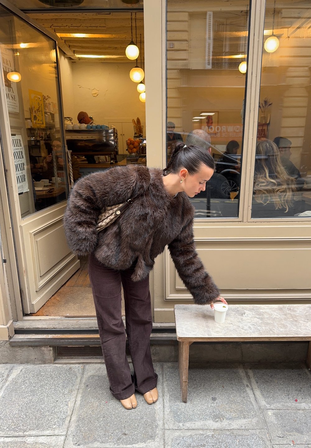 Chaqueta corta de piel sintética marrón, pantalones anchos marrones y zapatos planos beige. La persona alcanza una taza blanca sobre un banco de madera fuera de una cafetería.