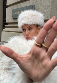 Witte jas van namaakbont met een bijpassende hoed, gouden ringen aan de vingers en een wazige achtergrond. De hand is voor het gezicht geheven.