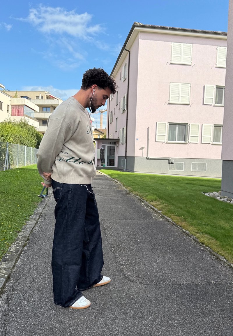 Homem com auriculares segura óculos de sol atrás das costas, em pé num caminho pavimentado perto de prédios residenciais sob um céu azul.