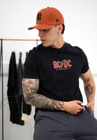 Crna pamučna majica s crvenim AC/DC grafikom, sivi šorts i narančasta bejzbol kapa s crnim logotipom. Tetovaže vidljive na ruci.