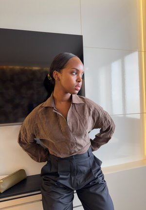 Mujer joven con el cabello recogido, vestida con una camisa marrón con cremallera y pantalones negros de talle alto, de pie en interiores cerca de una pantalla de televisión.