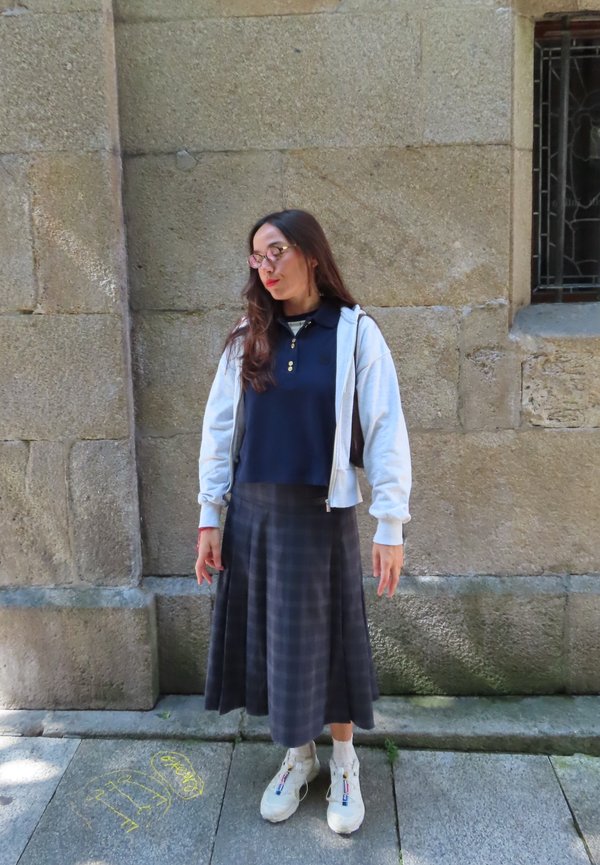 Felpa grigia con zip, polo blu navy e gonna lunga a quadretti. Sneakers bianche con accenti colorati. Capelli sciolti e occhiali. Sfondo di muro di pietra.