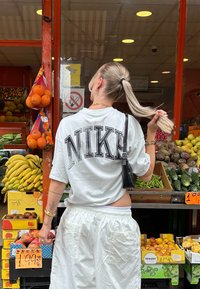 Fehér, rövid ujjú ing, fekete "NIKE" felirattal, laza fehér nadrággal párosítva; gyümölcsöket bemutató piaci háttér.