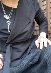 Μαύρη μπλούζα τύπου wrap με μακριά μανίκια και ζώνη. Σατινέ μαύρη φούστα με πτυχώσεις. Ασημένιο κολιέ με pendant σε σχήμα κλειδιού και κοχυλιού.