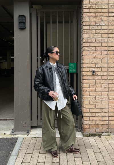 Persona con chaqueta de cuero negra, camisa blanca y pantalones verdes, sosteniendo una taza de café y una bolsa negra, de pie cerca de una pared de ladrillos y una puerta.