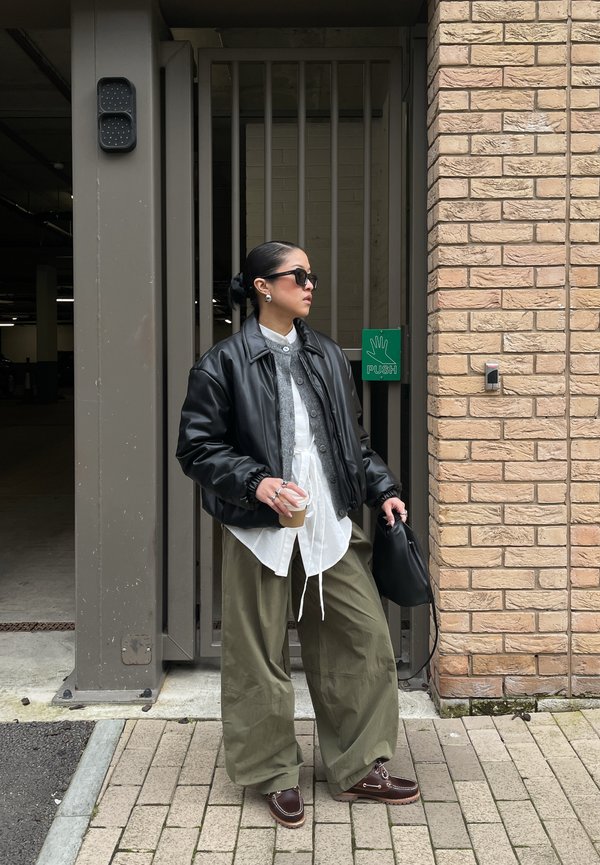 Person wearing a black leather jacket, white shirt, and green pants holding a coffee cup and black bag, standing near a brick wall and gate.