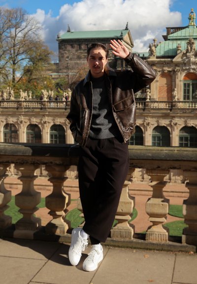 Modelo con una chaqueta de cuero marrón sobre un suéter gris, pantalones negros y zapatillas blancas, posando junto a una balaustrada de piedra con un fondo histórico.