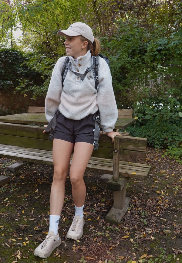 Fleece-genser i lys grå med knapper, kombinert med svarte shorts. Hvite sokker og lyse joggesko, utstyrt med en ryggsekk. Utendørs setting.