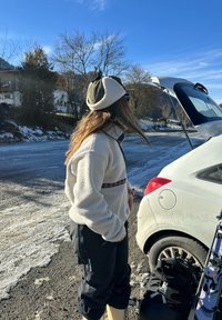 Veste en polaire crème avec une bande décorative, associée à un pantalon de ski noir. Bottes marron clair. Coffre de voiture ouvert avec skis et sac visibles.