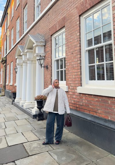 Chaqueta blanca acolchada sobre un suéter gris, jeans de pierna ancha azul, zapatos oscuros y un bolso burdeos, de pie junto a un edificio de ladrillo.