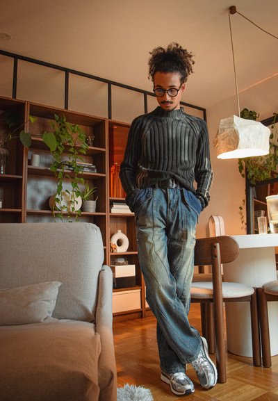 Hombre con gafas y cabello rizado, vestido con un suéter acanalado y jeans, apoyado en muebles de comedor en una sala acogedora llena de plantas.