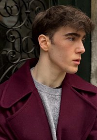 Manteau en laine bordeaux avec de larges revers porté sur un pull texturé en maille torsadée grise ; sujet aux cheveux courts et foncés et à la peau claire.