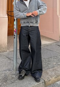 Light grey zip-up hoodie over a white shirt, paired with dark blue wide-leg jeans and black footwear with grey laces, standing by a pole.