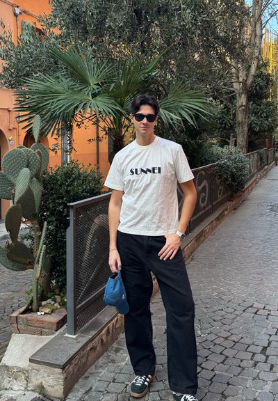 Una persona está de pie en un camino de adoquines, vistiendo una camiseta de algodón blanca con la palabra "SUNNEI" en negro, pantalones anchos negros y gafas de sol.