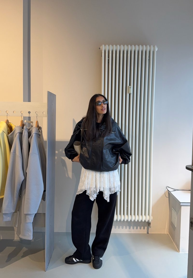 Casaco oversized em couro preto sobreposto a um top branco de renda, combinado com calças pretas e sapatos pretos, num ambiente interno moderno.