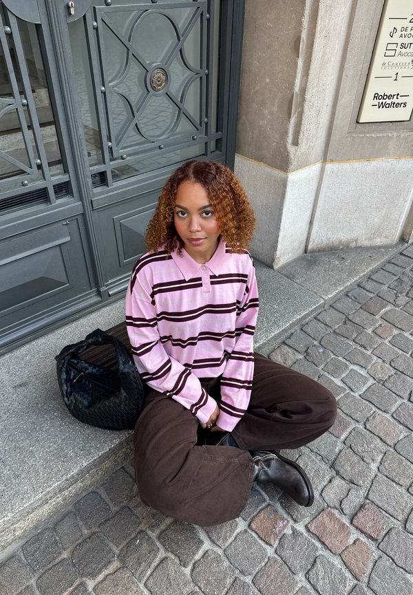 Young woman with curly hair wears pink striped shirt and brown pants sitting cross-legged on stone steps by metal door, black woven bag beside her.