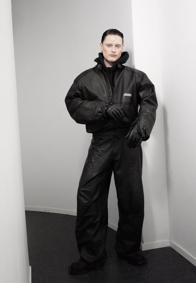 Persona con el cabello peinado hacia atrás, vistiendo una chaqueta de cuero negra de gran tamaño, pantalones y guantes, de pie en un pasillo blanco estrecho.