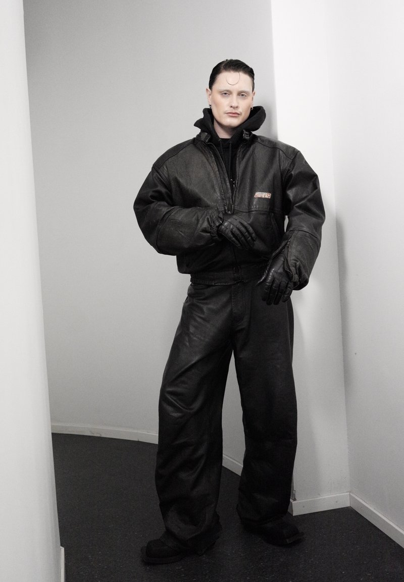 Personne aux cheveux tirés en arrière portant une veste en cuir noire surdimensionnée, un pantalon et des gants, debout dans un couloir blanc étroit.